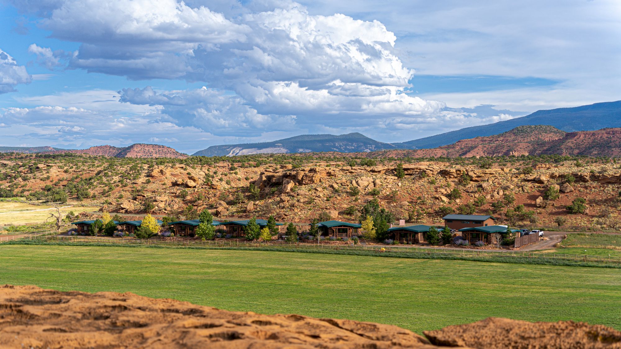 The image shows a desert landscape with greenery, buildings, and distant mountains under a cloudy sky.