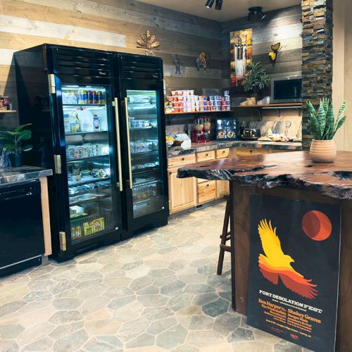 The image shows a cozy kitchen with a wooden theme, featuring fridges, shelves with snacks, plants, and a unique countertop with seating.
