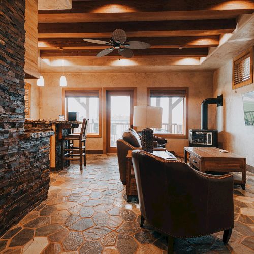 A cozy living area with stone flooring, wooden accents, seating, a mounted TV, and a kitchen island with chairs under pendant lights.