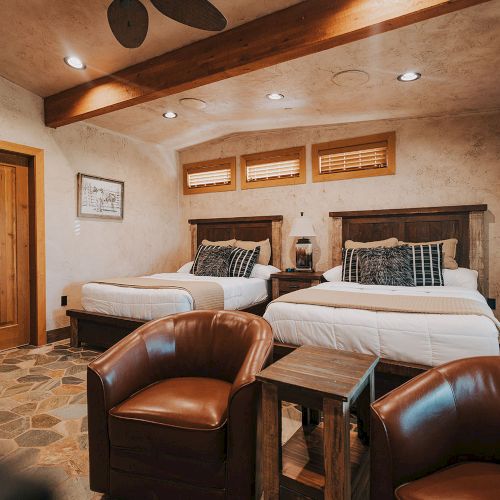 A cozy room with two beds, wooden furniture, leather chairs, and soft lighting creating a warm atmosphere.