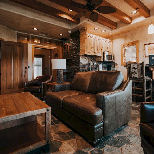 The image shows a cozy living room with leather furniture, a wooden coffee table, and a small kitchen area with overhead lighting and rustic decor.