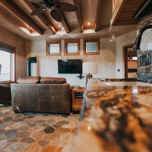 A cozy living room with a stone floor, brown sofa, wall-mounted TV, and visible kitchen counter, creating a warm and inviting atmosphere.