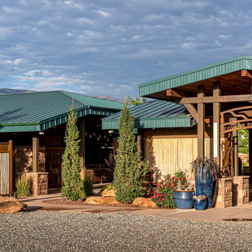 The image shows a rustic building with green roofs, wooden structures, and potted plants, set against a cloudy blue sky.