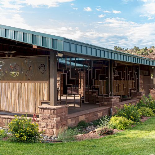 The image shows a modern building with metal and wood paneling, surrounded by landscaped gardens, under a partly cloudy sky.