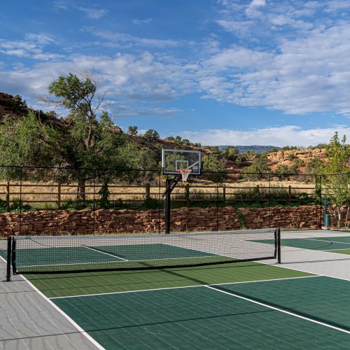 A multi-sport court with pickleball markings and a basketball hoop, set in a scenic outdoor area with trees and hills under a blue sky.