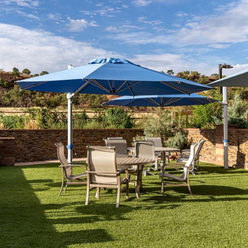 Outdoor patio with tables, chairs, and blue umbrellas on a grassy area, surrounded by a stone wall and scenic landscape.