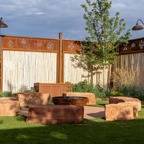 Outdoor setting with stone benches around a fire pit on grass, enclosed by a wooden fence with decorative motifs.