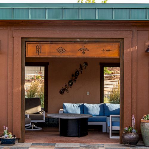 A small, outdoor lounge with a brown exterior, blue sofa, and decorative pots. The structure features wall designs and two exterior lights.