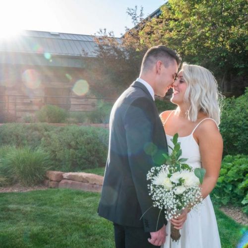 A couple in wedding attire stands together in a garden, the sun shining brightly behind them as they share an intimate moment.