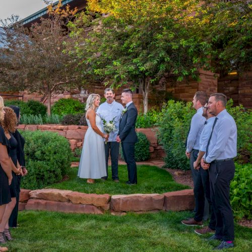 A wedding ceremony in a garden setting with a couple holding hands, surrounded by guests and officiant.
