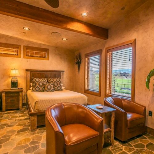 A cozy bedroom with a rustic design, featuring a queen bed, bunk bed, two chairs, a side table, and large windows overlooking greenery.