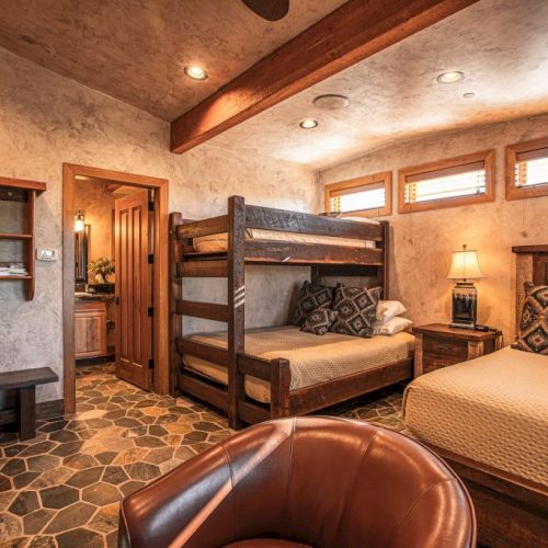 A cozy bedroom with a bunk bed, double bed, chair, and warm lighting. The floor is stone-tiled, with wooden accents throughout the room.