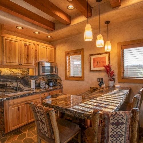 A rustic kitchen with wooden cabinets, stone accents, pendant lights, and a granite countertop with four upholstered chairs.