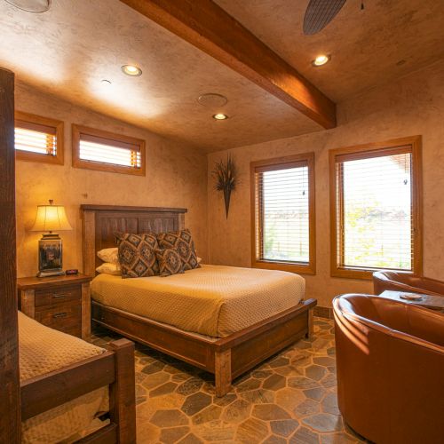 A cozy bedroom with a queen bed, bunk bed, chairs, wooden furniture, and warm lighting, featuring a plant and patterned floor.