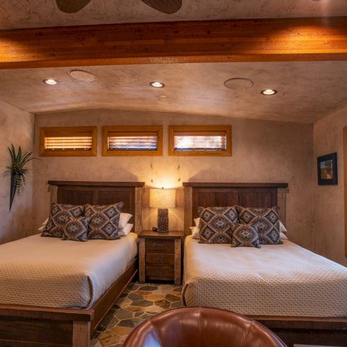 The image shows a cozy bedroom with two beds, a nightstand with a lamp, paintings on the walls, and a window view.