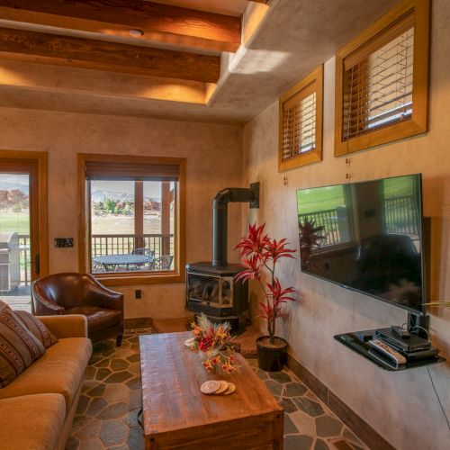A cozy living room features a TV, sofa, plants, wood stove, and a view of the outdoors through windows and a glass door.