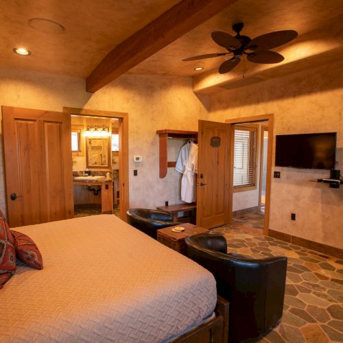 A cozy bedroom features a bed, armchairs, a TV, wooden accents, a ceiling fan, unique flooring, and an accessible bathroom.