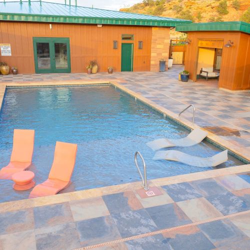 A swimming pool with two pink and two gray loungers on the water, surrounded by tiled flooring and wooden buildings outdoors.