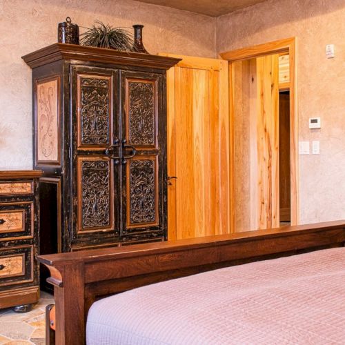 A cozy bedroom features a wooden bed, ornate wardrobe and dresser, a TV, and flower arrangements, with warm lighting throughout.