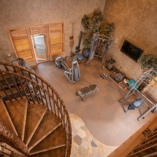 The image shows a gym with exercise equipment, a TV, and plants, viewed from a spiral staircase.