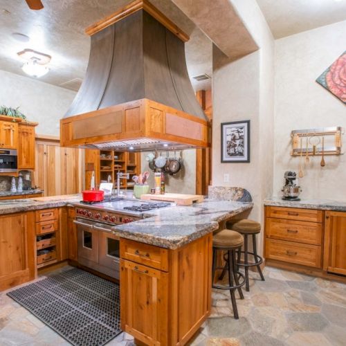 A spacious kitchen with wooden cabinets, a central island with stools, and a large range hood. Art decorates the walls.