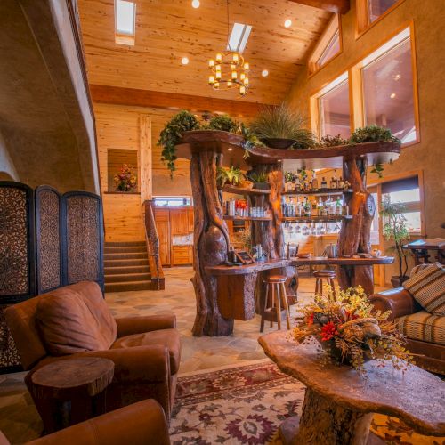 A cozy rustic living room with wooden furniture, plants, a bar area, and warm lighting, creating an inviting, relaxed atmosphere.