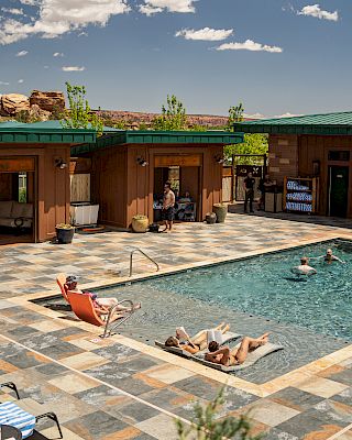 A pool surrounded by people and buildings under a sunny sky; some are swimming, others relax on loungers or walk around the deck.