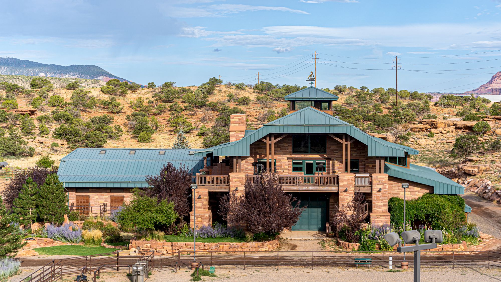 A large, rustic house with a green roof is set in a desert landscape, featuring stone walls and surrounded by vegetation.