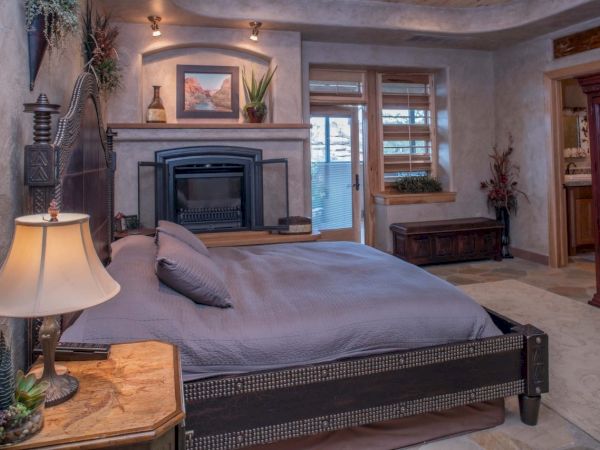A cozy bedroom with a large bed, fireplace, plants, and a wooden cabinet, featuring warm lighting and a decorative ceiling.