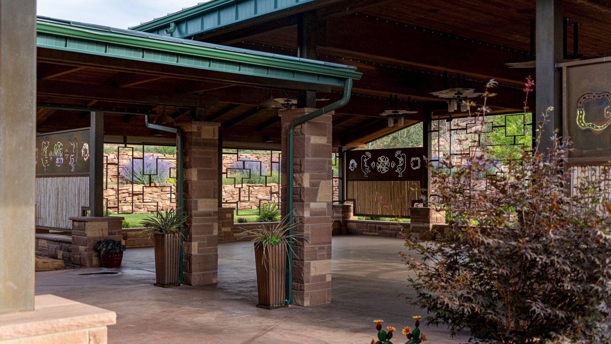 The image shows a covered outdoor space with stone pillars, greenery, and wall art, resembling a patio or gathering area.