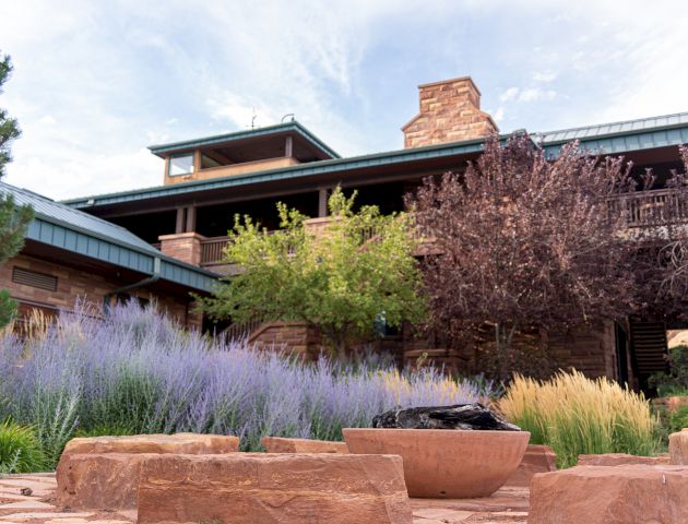 A rustic building with a stone and wood exterior surrounded by lush landscaping and a fire pit area in the foreground.