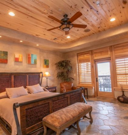 A cozy bedroom with a wooden ceiling, stone floor, fireplace, and large bed, featuring warm lighting and a rustic decor theme.