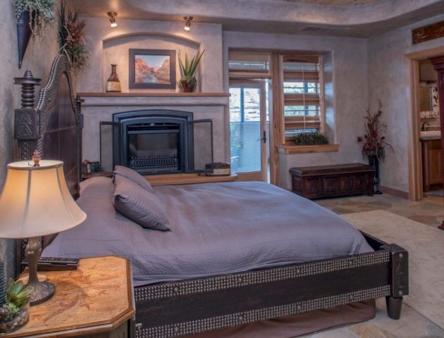 A cozy bedroom with a large bed, fireplace, plants, and a window with wooden blinds, creating a warm and inviting atmosphere.