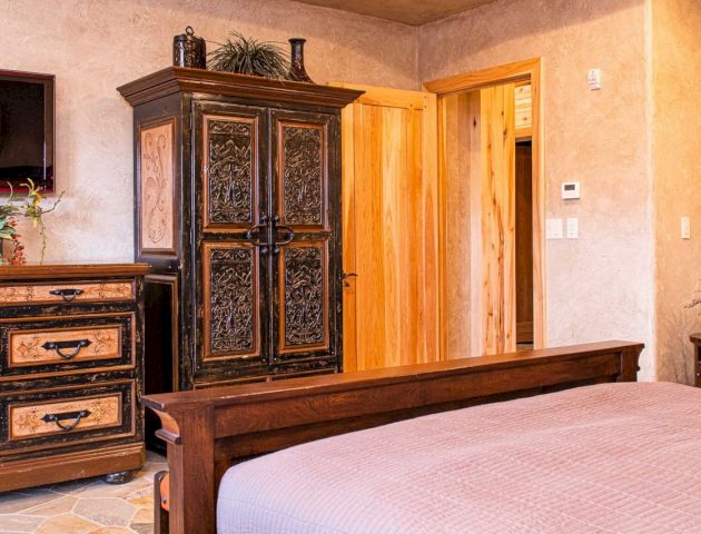 A cozy bedroom features a wooden bed, ornate wardrobe, dresser, wall-mounted TV, and floral decor, with warm lighting enhancing the ambiance.