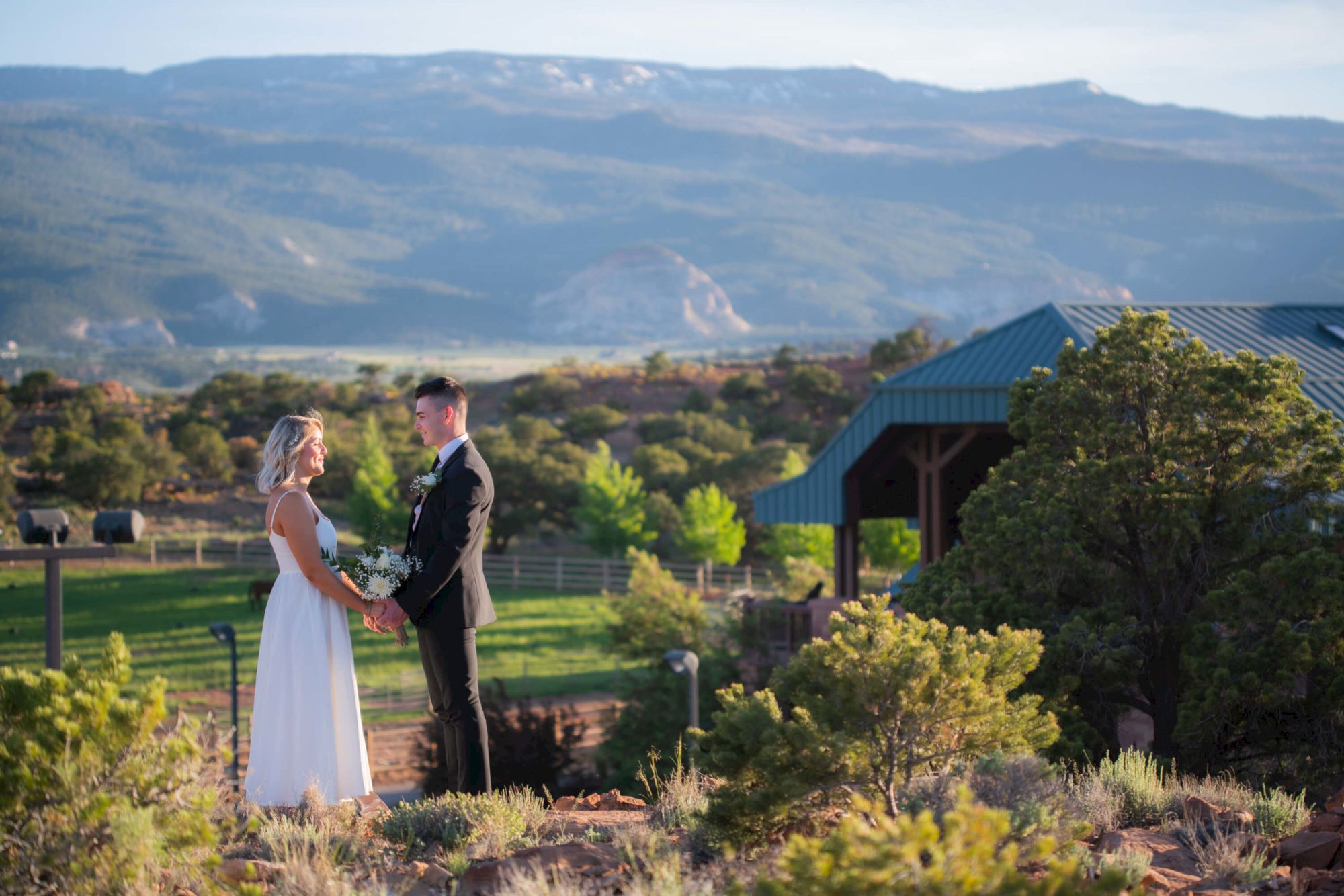 A couple in wedding attire stands outdoors amid scenic mountains and greenery, with a gazebo in the background.