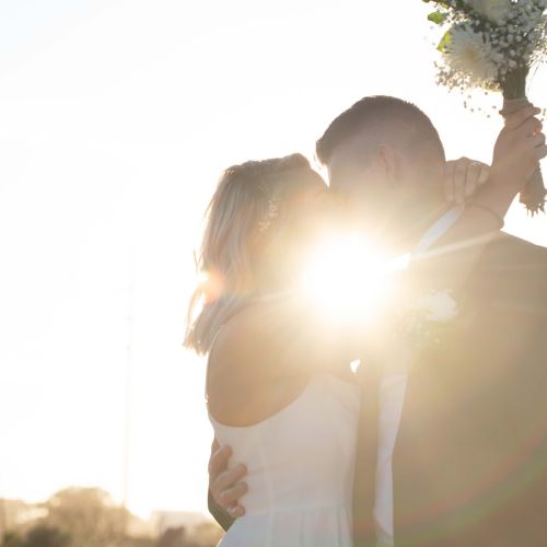 A couple embraces and kisses with the sun shining behind them, holding a bouquet aloft against a bright sky.