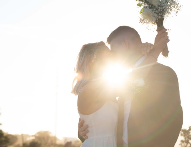 A couple embraces and kisses with the sun shining behind them, holding a bouquet aloft against a bright sky.