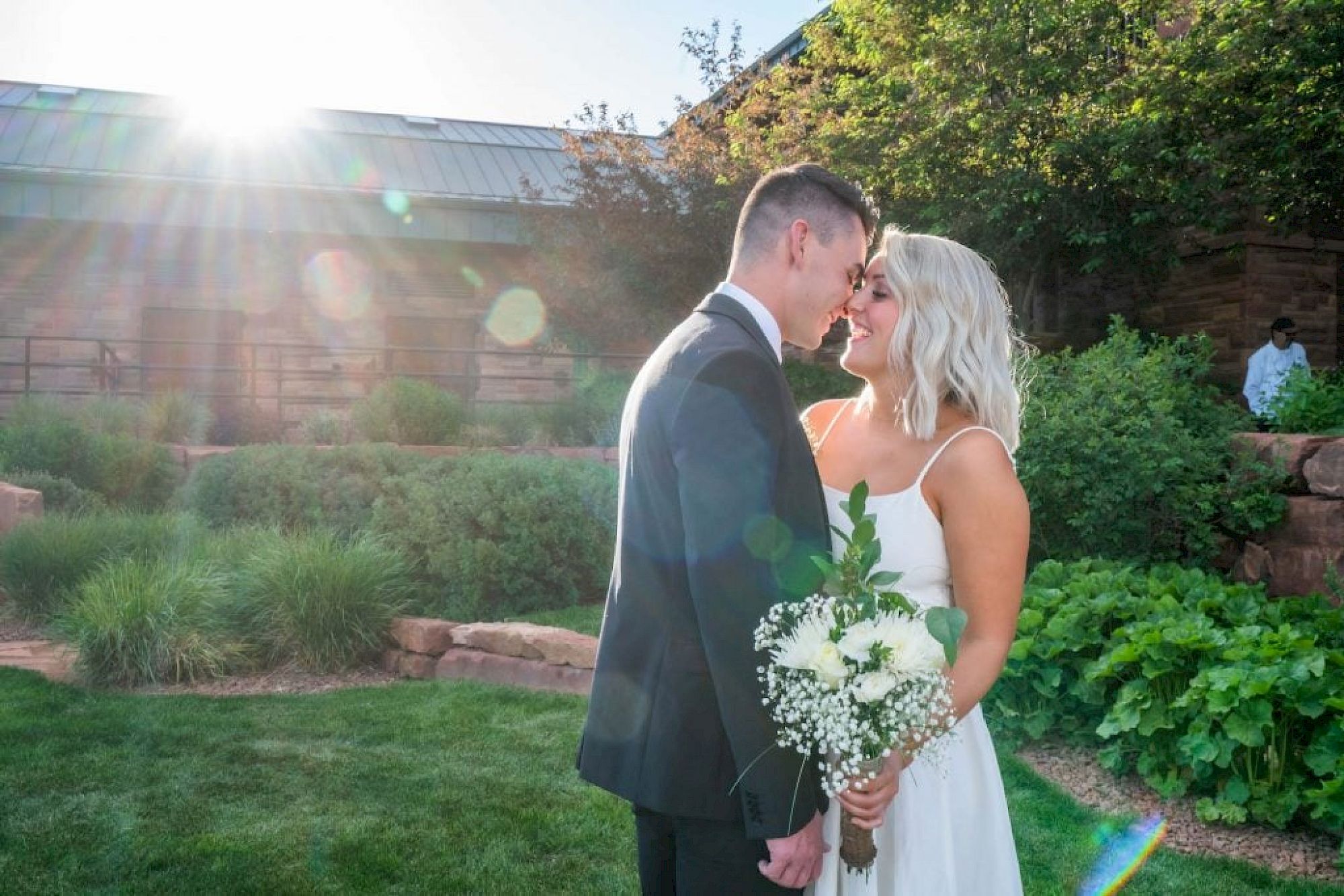 A couple in wedding attire stands close, smiling at each other in a garden with sunlight filtering in from the top left.