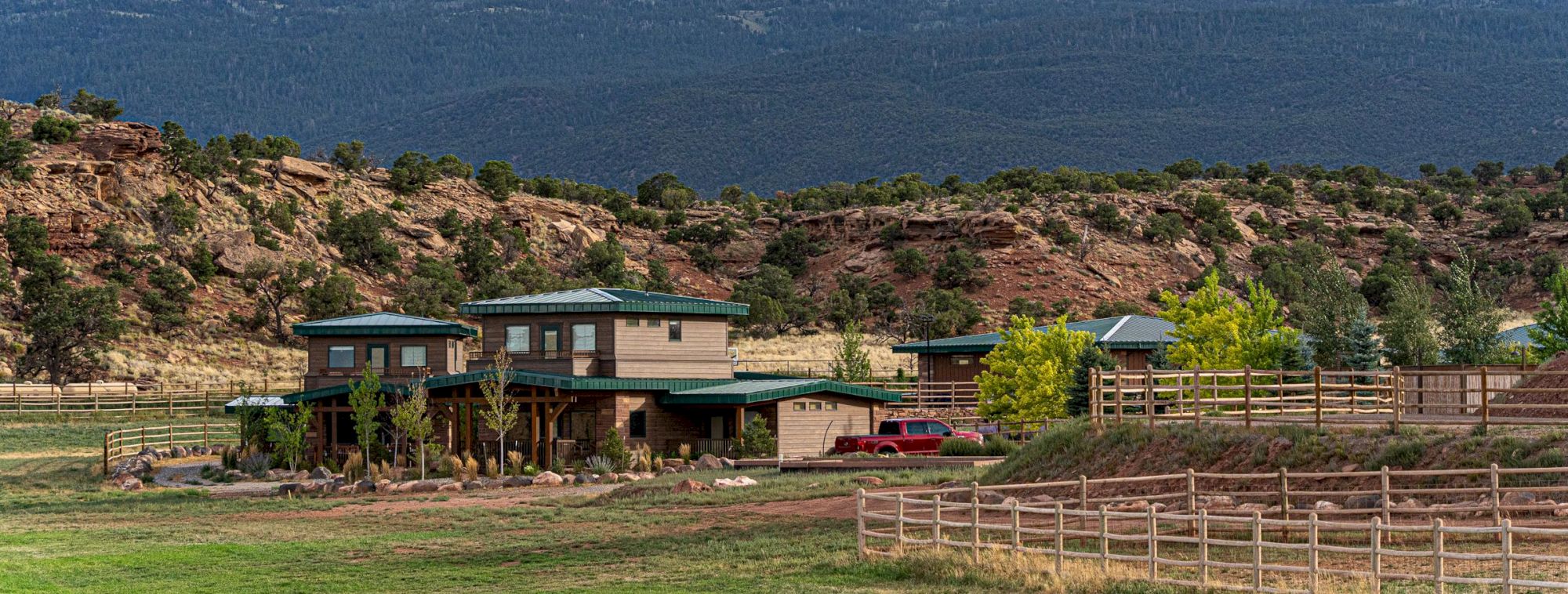 A ranch-style house with green roofs surrounded by trees, fences, and mountains under a partly cloudy sky completes the scene.