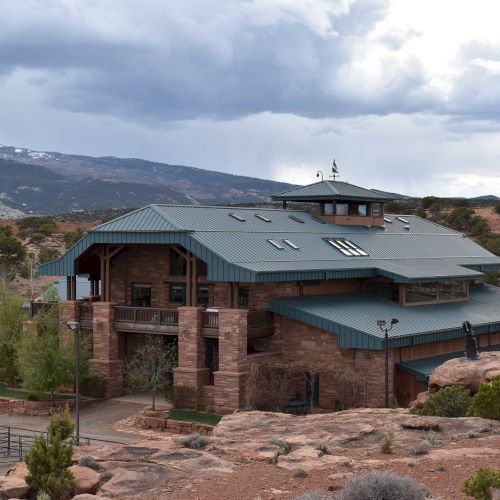The image shows a large, rustic building with a green roof set in a desert landscape with mountains and cloudy skies in the background.