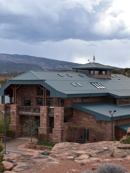 The image shows a large, rustic building with a green roof set in a desert landscape with mountains and cloudy skies in the background.