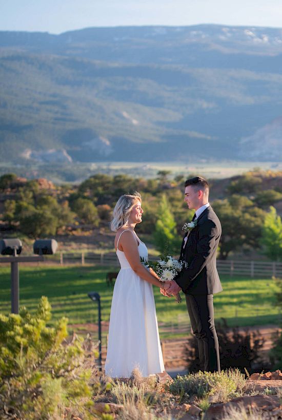 A couple in a wedding setting stands outdoors, holding hands, with mountains and greenery in the background.