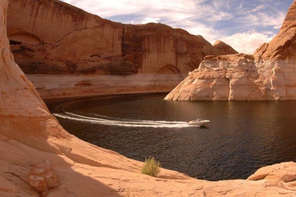 A speedboat cruises along a winding waterway surrounded by towering sandstone cliffs under a partly cloudy sky.