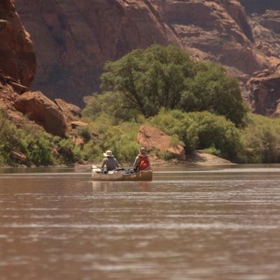Two people in a canoe are paddling on a calm river surrounded by rocky cliffs and greenery.