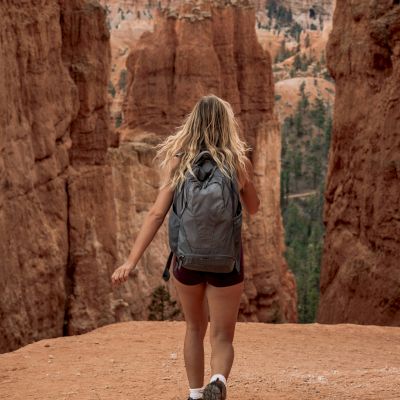 A person with a backpack walks towards stunning red rock formations, possibly in a canyon. The scene is scenic and adventurous.