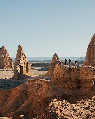 A group of people stand on a rocky outcrop, surrounded by dramatic sandstone formations in a desert landscape under a clear sky.