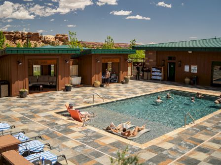 A poolside scene with a few people relaxing, clear skies, and buildings in the background, set in a tiled area.