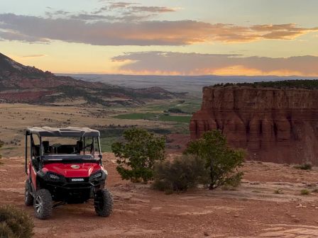 A red off-road vehicle is parked on a rocky terrain with a scenic landscape of cliffs, trees, and a sunset in the background.