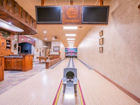 The image shows a home bowling alley with wooden interiors, two screens, and a bar area to the side.