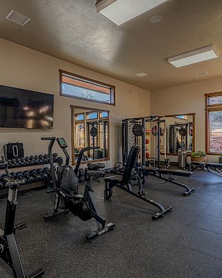 The image shows a gym with exercise bikes, weight benches, and free weights, along with large windows and a TV on the wall.